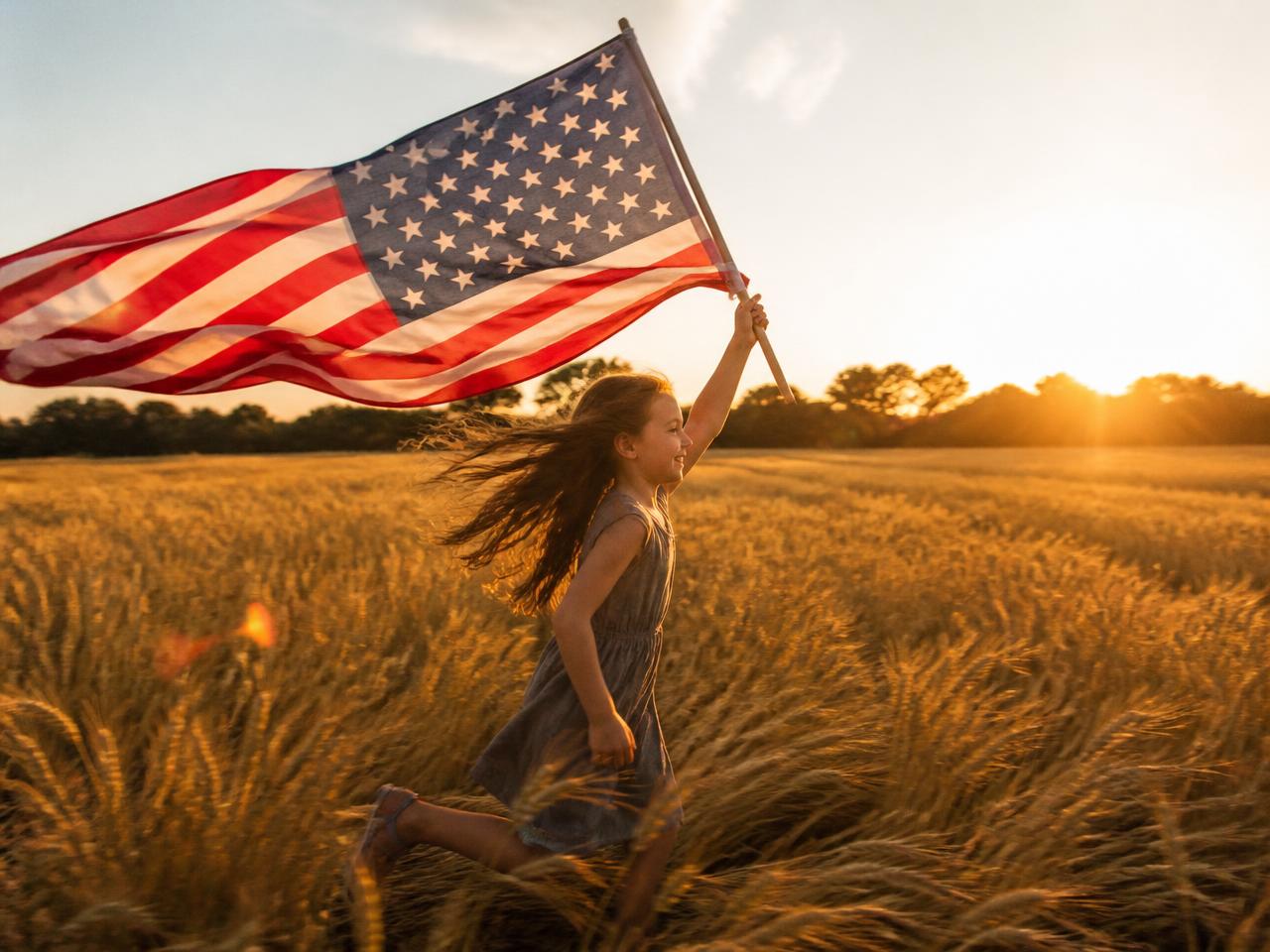 A child running through a sunlit wheat field carrying an American flag